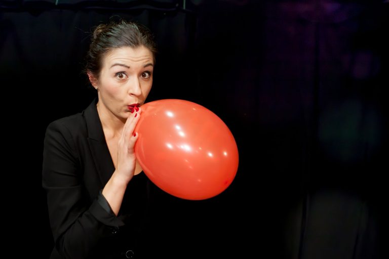 Kobieta w czarnej marynarce nadmuchuje czerwony balon, wyglądając na zaskoczoną, jakby przygotowywała się do karnawału, na ciemnym tle.