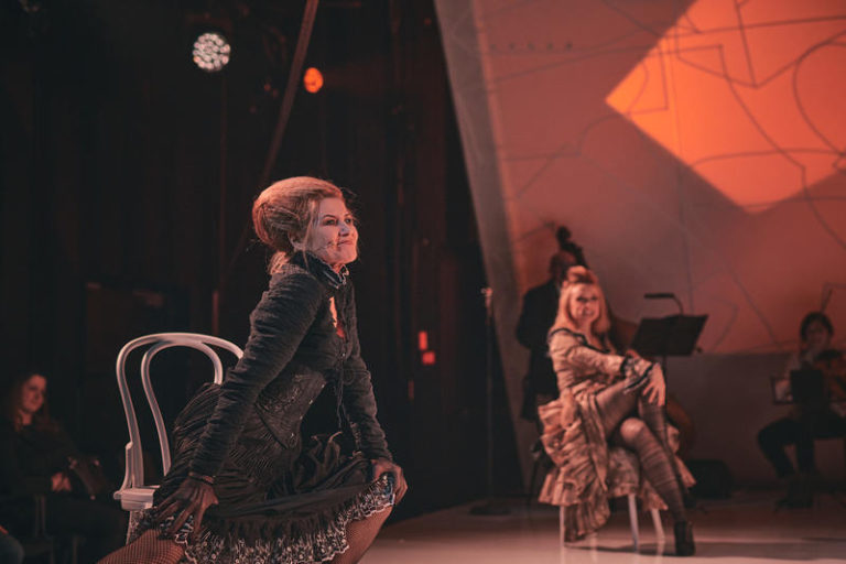 Zdjęcie ze spektaklu teatralnego. Na scenie w swobodnych pozach siedzą dwie aktorki. Ubrane są w długie suknie z falbanami. Włosy mają upięte w wysokie koki. Z tyłu po prawej stronie mężczyzna gra na kontrabasie, a kobieta na skrzypcach. Aparat ustawiony jest tak, iż kadr obejmuje również część publiczności.