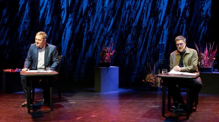 Dwóch mężczyzn siedzi przy osobnych stołach na słabo oświetlonej scenie z niebieskimi smugami światła i kompozycjami kwiatowymi za nimi. Obaj mają mikrofony i dokumenty, co sugeruje dyskusję lub występ.