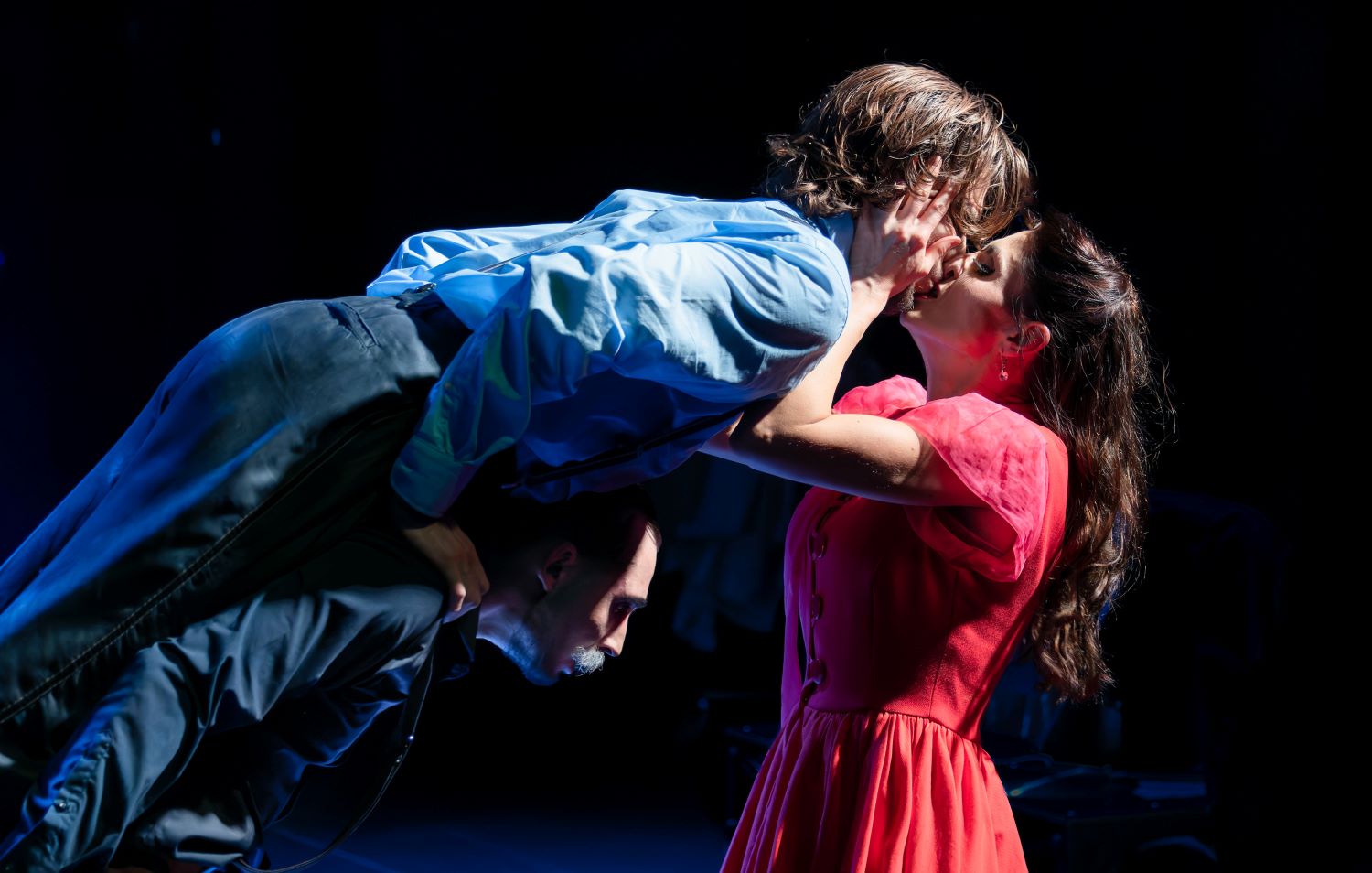 Kobieta w czerwonej sukience namiętnie całuje osobę w niebieskiej, która jest trzymana poziomo przez innego mężczyznę - intensywna scena, dramatycznie oświetlona, przywołująca energię i romans balu o północy na ciemnym tle.