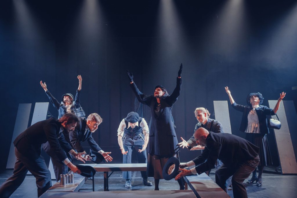 Grupa aktorów w dramatycznych pozach na słabo oświetlonej scenie wywołuje intensywne emocje, oddając ducha spektaklu Jerzego Zonia. Minimalistyczna scenografia i ciemne kostiumy dodają teatralnej atmosfery przypominającej Arkadię w teatrze.
