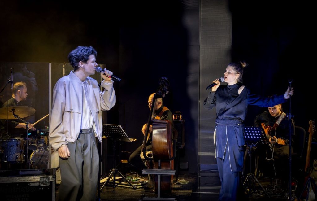 Dwóch wokalistów Teatru KTO występuje na scenie z mikrofonami, naprzeciwko siebie, podczas gdy zespół z perkusistą, basistą i gitarzystą gra w tle przy dynamicznym oświetleniu scenicznym.