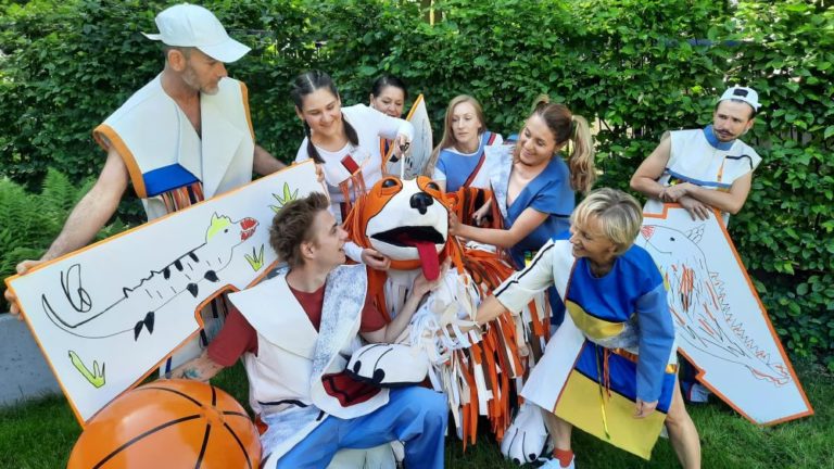 Grupa ludzi w kolorowych, zabawnych kostiumach, w tym dużych zwierząt i rekwizytów do koszykówki, gromadzi się wokół kogoś w dużym pomarańczowo-białym kostiumie psa, wszyscy uśmiechają się i wchodzą w interakcje na świeżym powietrzu z zielenią za nimi.