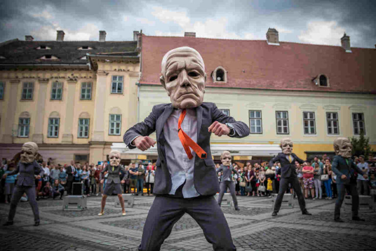 Grupa performerów w garniturach i przerośniętych maskach starców tańczy na brukowanym placu, z dużym tłumem obserwującym i zabytkowymi budynkami w tle.
