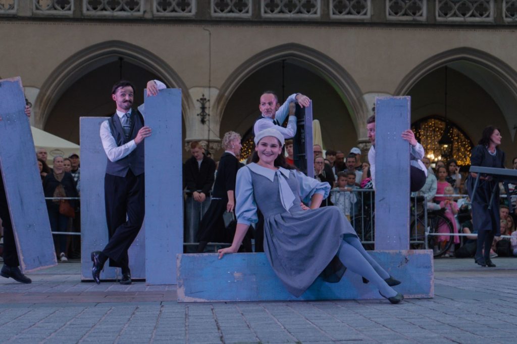 Grupa wykonawców Teatru KTO w kostiumach w stylu vintage pozuje z dużymi niebieskimi drewnianymi deskami podczas ulicznego spektaklu na świeżym powietrzu, z publicznością i zabytkowym budynkiem w tle. Kobieta w centrum siedzi i uśmiecha się do kamery.