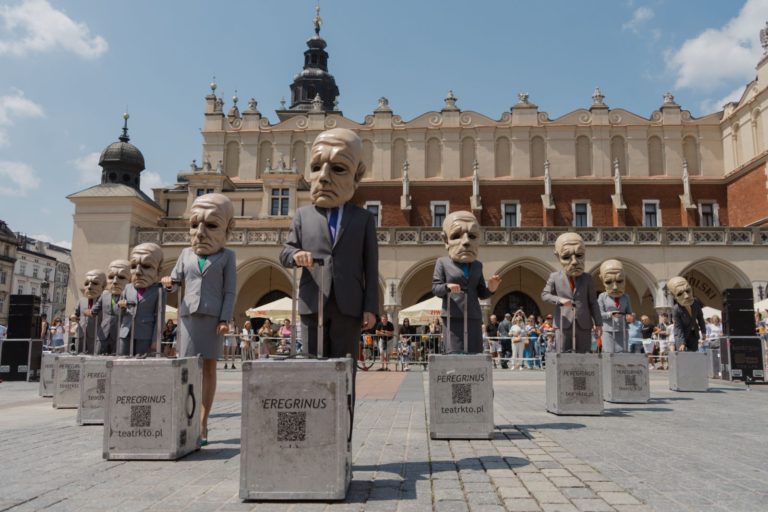 Performerzy z Teatru KTO, ubrani w obszerne garnitury i maski na głowach, stoją na betonowych blokach na miejskim placu. Zabytkowy budynek i tłumy w tle sugerują, że jest to część tętniącego życiem spektaklu ulicznego.