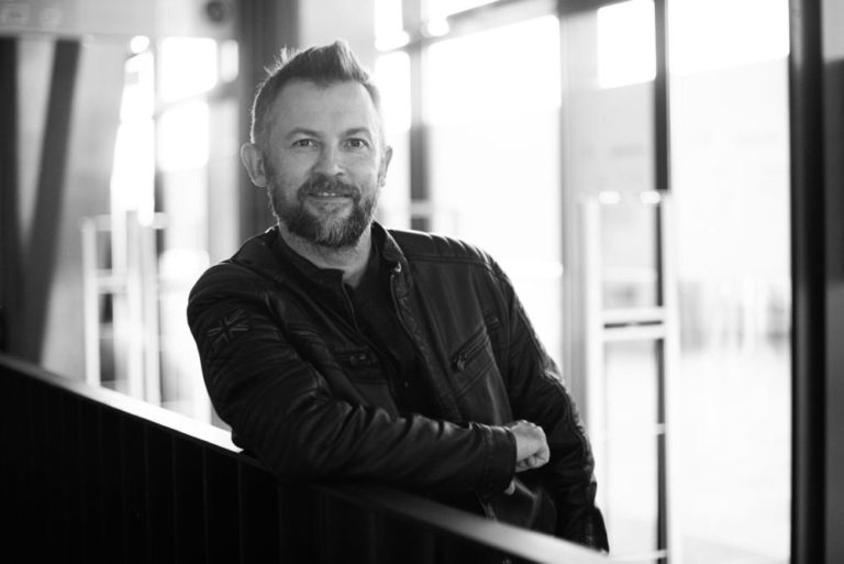 Mężczyzna z brodą i krótkimi włosami, ubrany w ciemną skórzaną kurtkę, opiera się o poręcz i uśmiecha się do kamery w jasnym, nowoczesnym wnętrzu. Obraz jest czarno-biały.