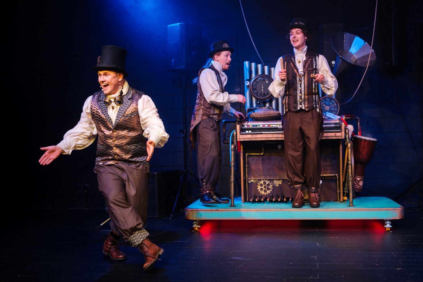 Trzech aktorów w kostiumach w stylu wiktoriańskim występuje na scenie; jeden tańczy z przodu, podczas gdy dwóch stoi za maszyną z rurami i pokrętłami, ustawioną na niebiesko oświetlonym tle sceny teatralnej.