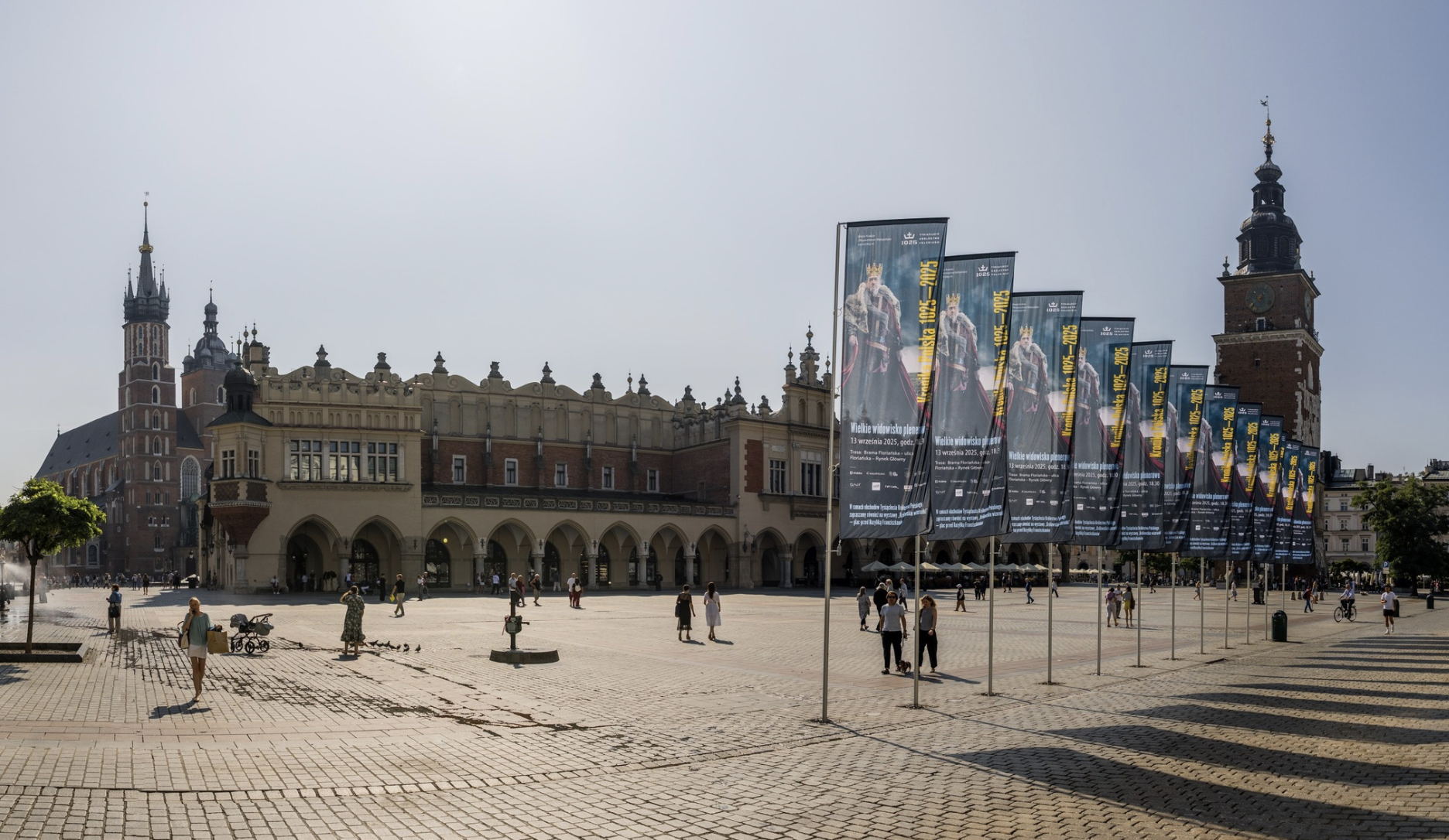 Ludzie spacerują po krakowskim Rynku Głównym, z Sukiennicami w centrum i Bazyliką Mariacką po lewej stronie, przypominając sceny opisane przez Pawła Głowackiego w jego felietonie "A nam którzyśmy w drodze". Kolorowe transparenty po prawej stronie pod czystym niebem.