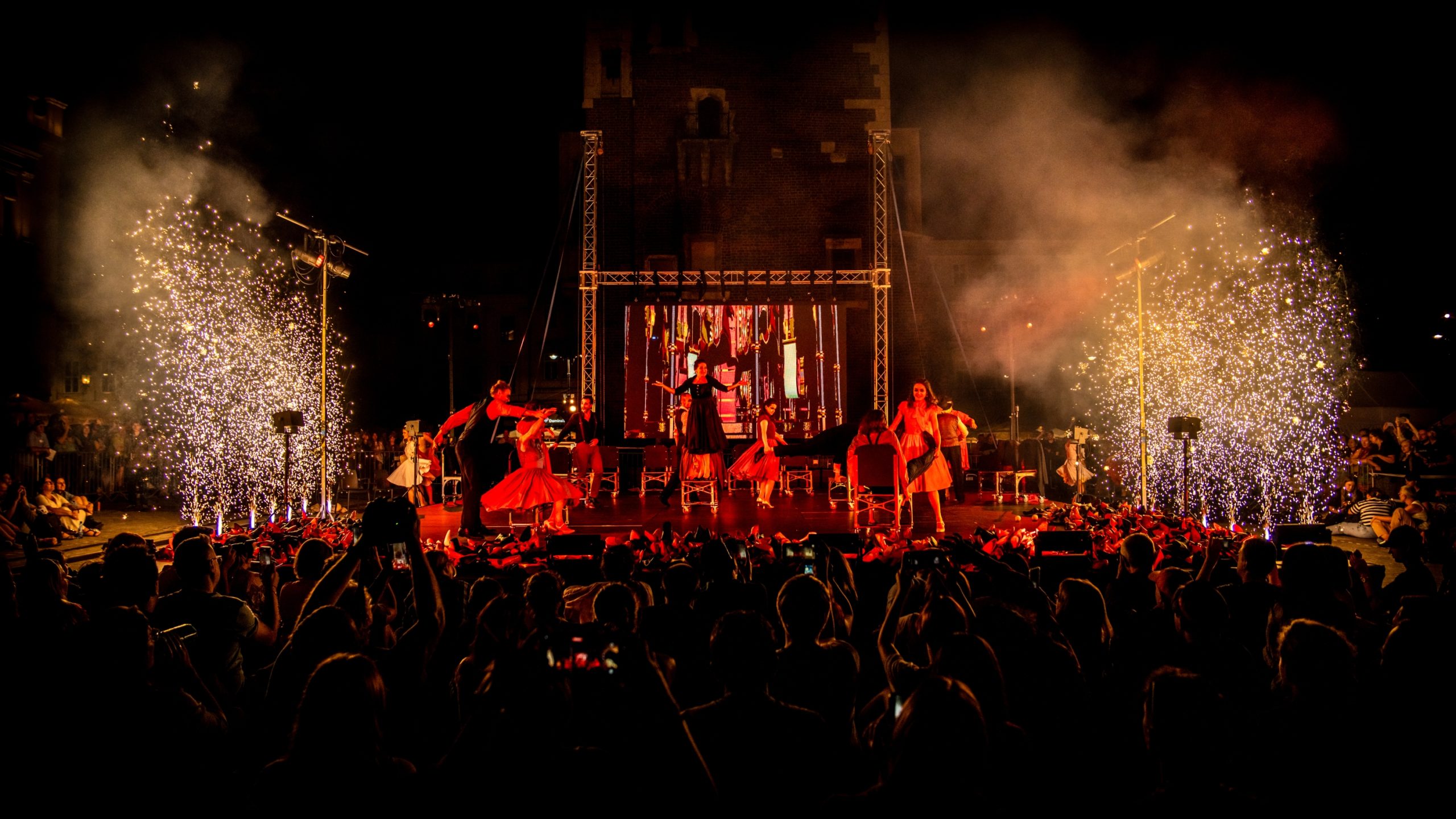 Tętniący życiem występ sceniczny w nocy w Londynie przedstawia tancerzy w żywych kostiumach i dramatycznym oświetleniu, z iskrami latającymi po obu stronach sceny, podczas gdy duży tłum obserwuje i robi zdjęcia. Teatr to wolność ożywiona.