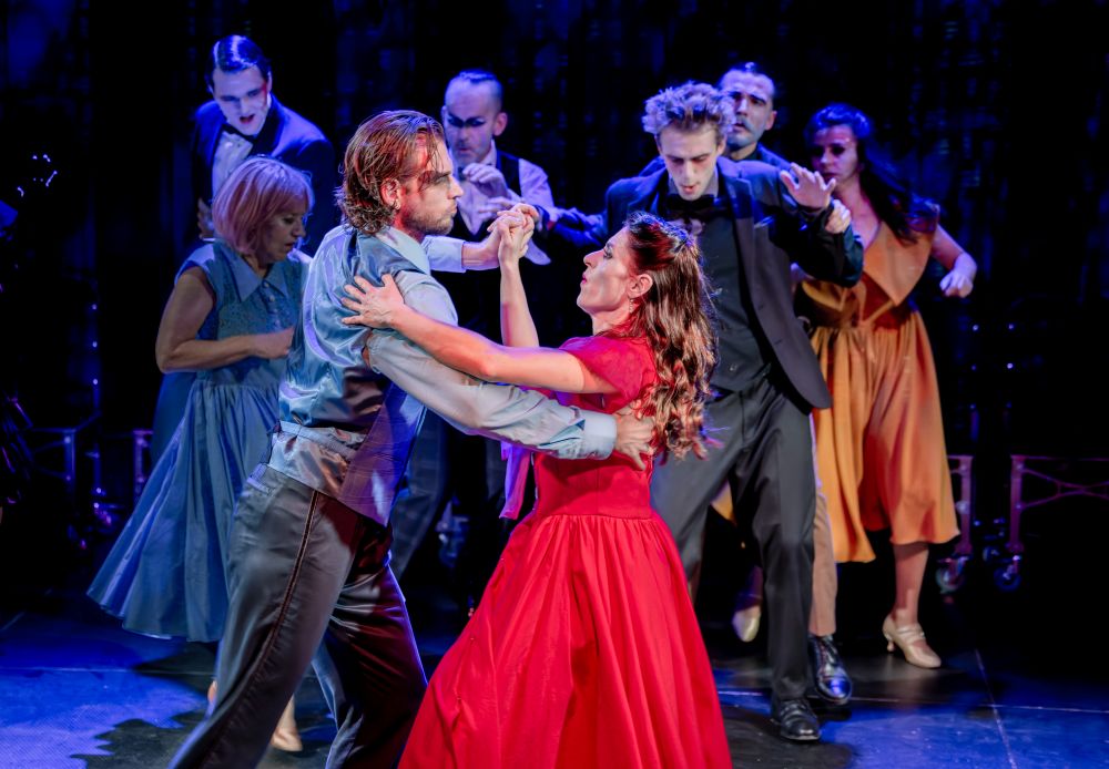 Mężczyzna i kobieta tańczą blisko siebie na pierwszym planie, oboje ubrani formalnie; inni w dramatycznych pozach i kostiumach stoją za nimi na scenie, przywołując świątecznego ducha uroczystości andrzejkowej lub przedstawienia teatralnego.