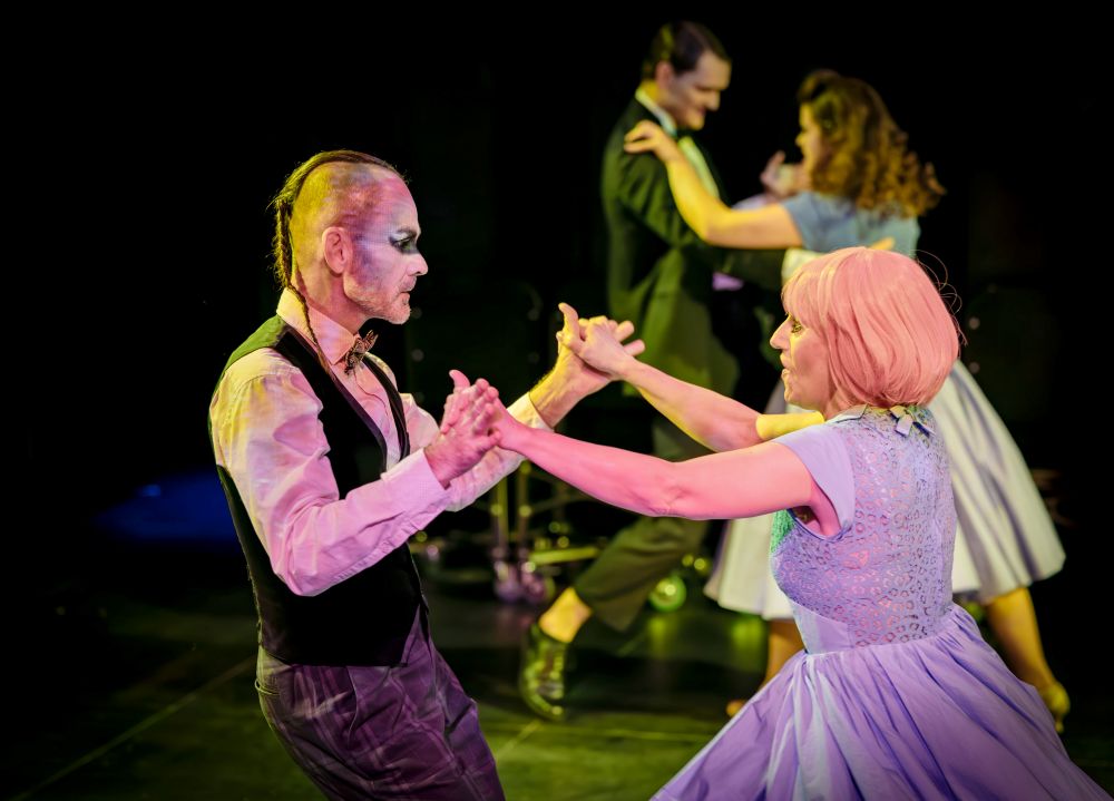 Dwie pary tancerzy występują na scenie, świętując andrzejki. Para z przodu obejmuje osobę z dramatycznym makijażem i kamizelką trzymającą się za ręce z osobą w jasnofioletowej sukience. Inna para tańczy w tle, nieco nieostro.