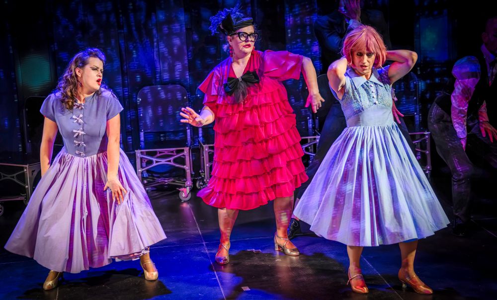 Trzy performerki w sukienkach w stylu vintage tańczą na scenie, ożywiając andrzejkową uroczystość. Dwie z nich ubrane są w jasnoniebieskie sukienki ze złotymi butami, podczas gdy środkowa olśniewa jasnoróżową sukienką, czarnymi okularami i nakryciem głowy na żywym tle.