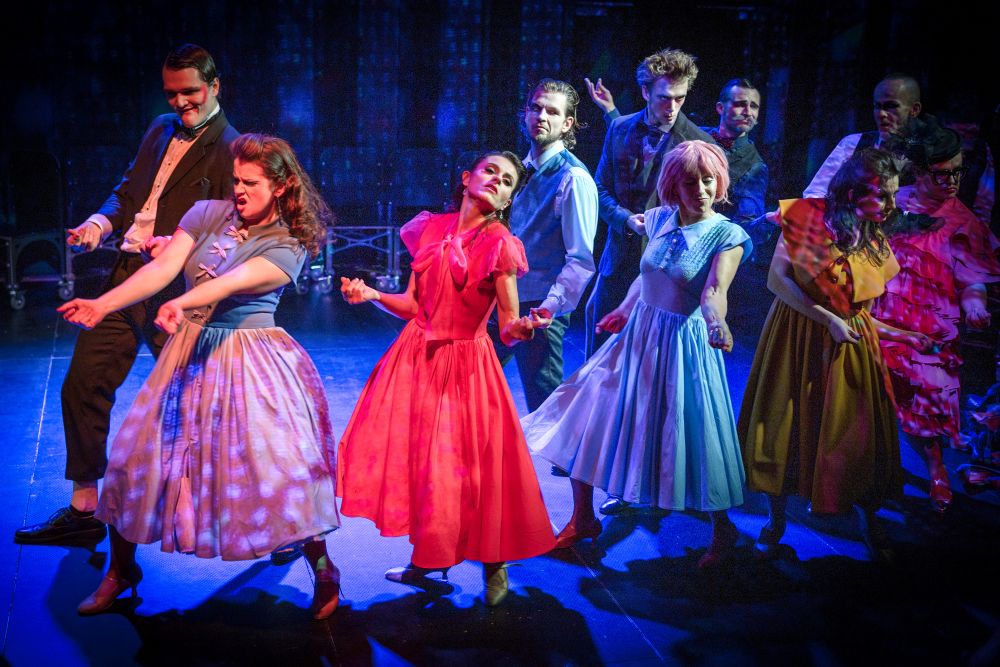 Grupa aktorów w kolorowych strojach vintage tańczy energicznie na scenie w dramatycznym oświetleniu, tworząc żywą i teatralną scenę andrzejkową.