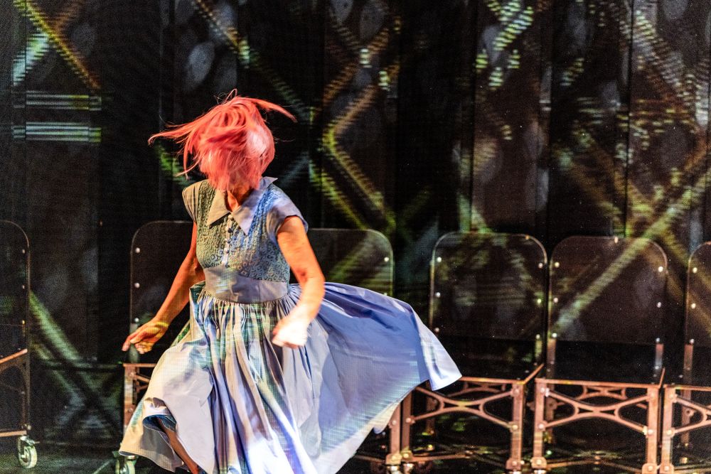 Osoba w jasnoniebieskiej sukience z różowymi włosami energicznie tańczy na scenie, świętując andrzejki, a w tle widać rzędy pustych krzeseł i wzorzyste oświetlenie.