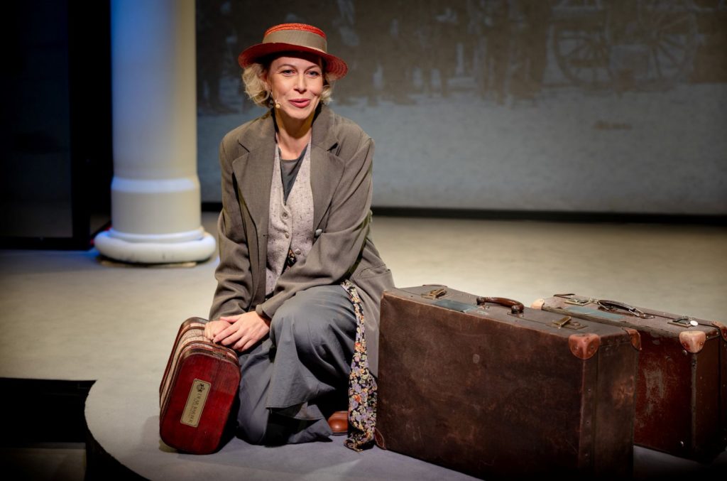 Kobieta w ubraniu vintage i czerwonym kapeluszu klęczy na scenie obok dwóch starych walizek, patrząc nieco poza kamerę, z neutralnym tłem i dużą białą kolumną za nią.