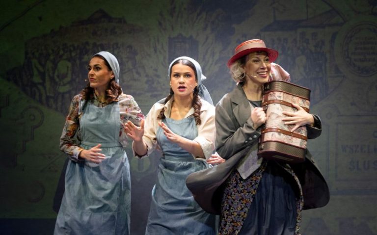 Trzy kobiety na scenie, dwie w niebieskich fartuchach i chustach na głowach, wyglądające na zaniepokojone, a jedna w kapeluszu i płaszczu uśmiechająca się, trzymając duży cylindryczny pakiet, z żółto oświetlonym tłem za nimi.