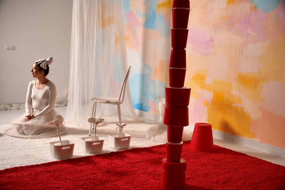 Kobieta w białym stroju i chustce na głowie siedzi na białym dywanie, patrząc w lewo. Za nią znajduje się białe krzesło, przezroczysta tkanina i pastelowa fototapeta. Na pierwszym planie na czerwonym dywanie ułożone są czerwone wiadra, a obok nich białe wiadra i sztuczne kwiaty.