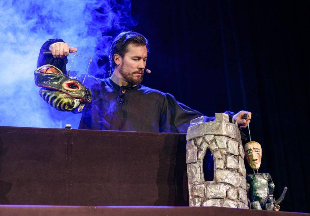 Lalkarz w ciemnym ubraniu wykonuje BAŚŃ O RYCERZU BEZ KONIA, zręcznie operując lalką smoka i lalką rycerza w pobliżu kamiennej wieży na scenie, z niebieskim dymem wirującym w tle.