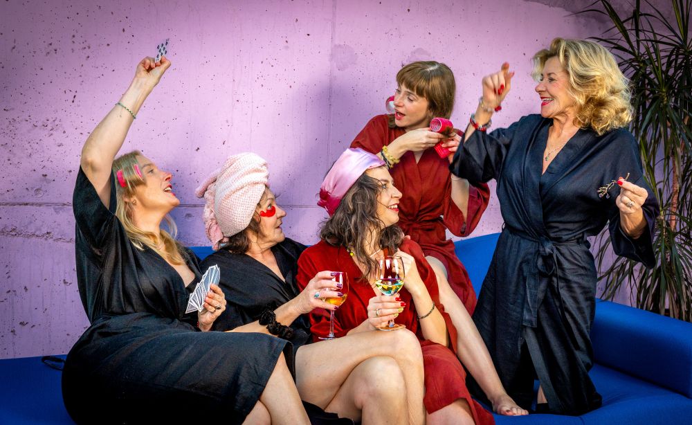 Pięć kobiet w szatach siedzi na niebieskiej kanapie przy różowej ścianie, śmiejąc się i świętując. Niektóre trzymają kieliszki z winem, jedna ma ręcznik na głowie i wydaje się, że cieszą się zabawnym, żywym spotkaniem.