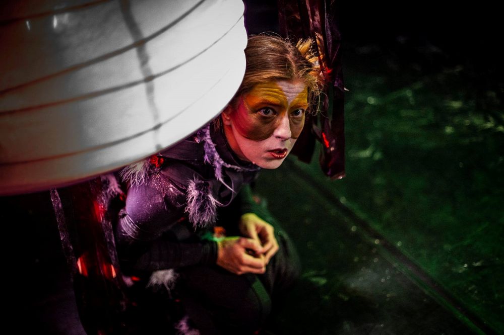 Performer w teatralnym makijażu i kostiumie, przypominający pająka, przykuca pod dużą strukturą przypominającą sieć na słabo oświetlonej scenie - część hipnotyzującego spektaklu Między oWadami - z dramatyczną czerwoną i żółtą farbą na twarzy i futrzanymi detalami.