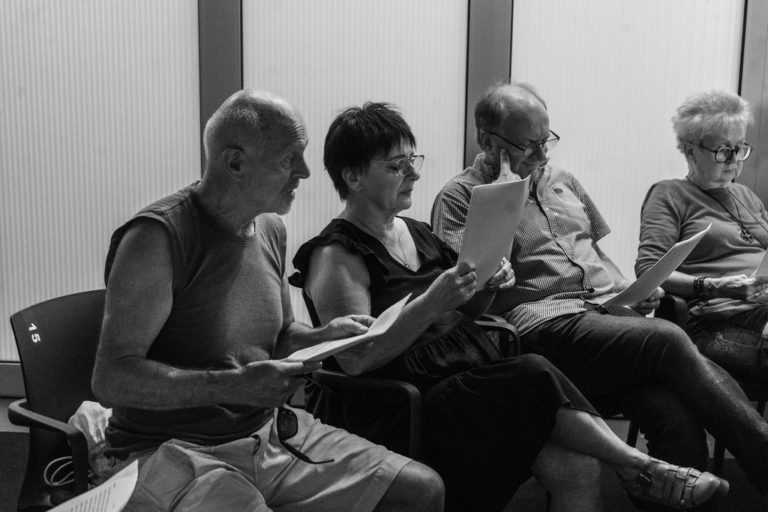 Cztery starsze osoby siedzą na krzesłach w pomieszczeniu, uważnie czytając lub śpiewając z wydrukowanych kartek. Obraz jest czarno-biały, podkreślając ich skupione miny - być może dzielą się wspomnieniami z października lub wskazówkami na temat sadzenia tulipanów - w relaksującym otoczeniu.