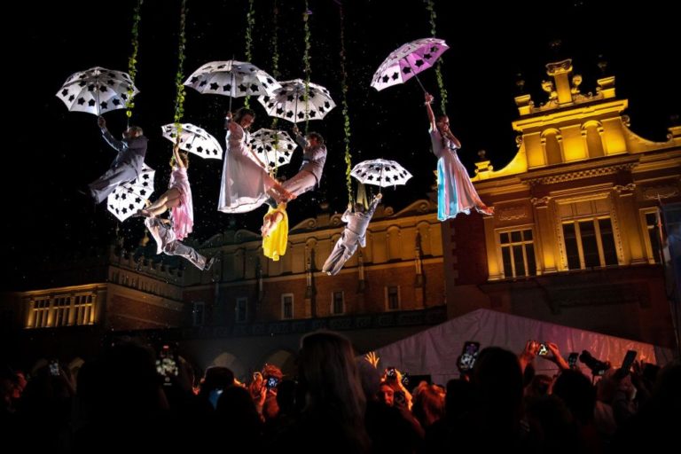 Performerzy w białych kostiumach z Teatru KTO zwisają z otwartymi parasolami nad nocnym tłumem. Oświetlony zabytkowy budynek wzmacnia intensywność sceny, gdy widzowie oglądają i rejestrują pokaz, podziwiając teatralną odwagę.