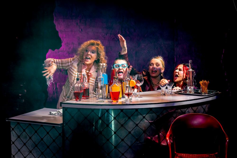 Cztery osoby siedzą przy barze z różnymi butelkami i kieliszkami, śmiejąc się i wykonując animowane gesty. Żywe, kolorowe oświetlenie odzwierciedla poczucie wspólnoty i teatralnej odwagi, przypominając żywego ducha Teatru KTO.