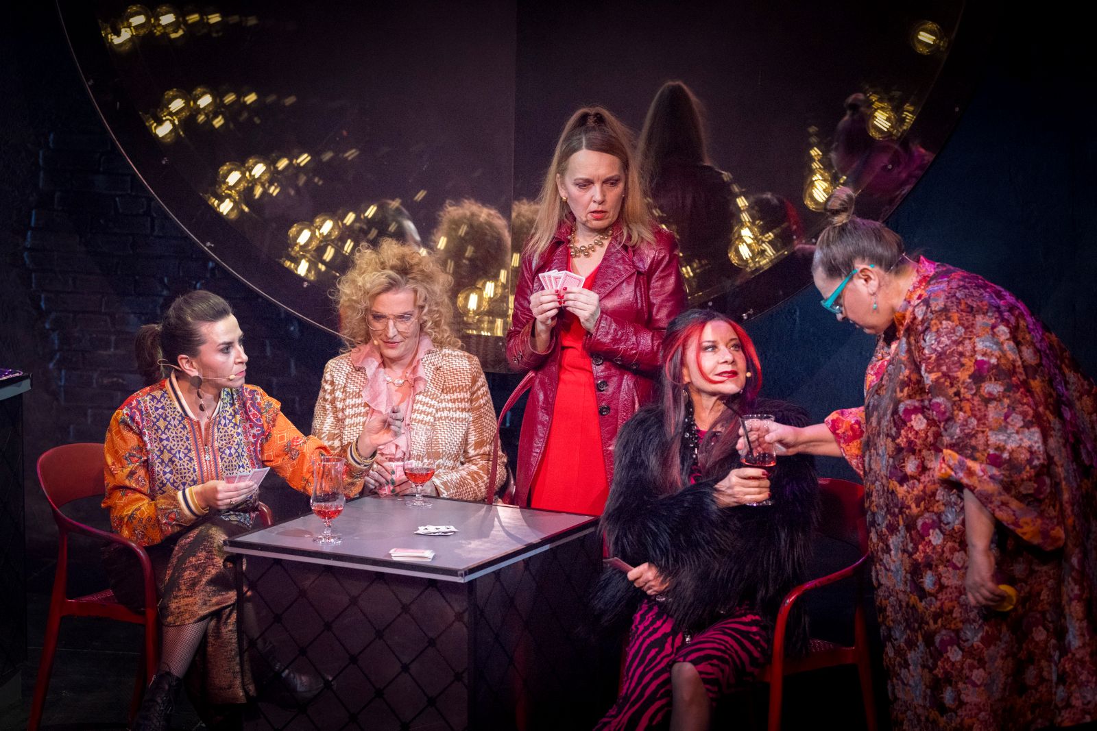 Pięć osób w kolorowych, eklektycznych strojach gromadzi się wokół małego stolika, grając w karty, a ich gesty i mimika są pełne dramatyzmu - jak scena prosto z Salonu Bizarre w tętniącej życiem scenerii teatralnej.