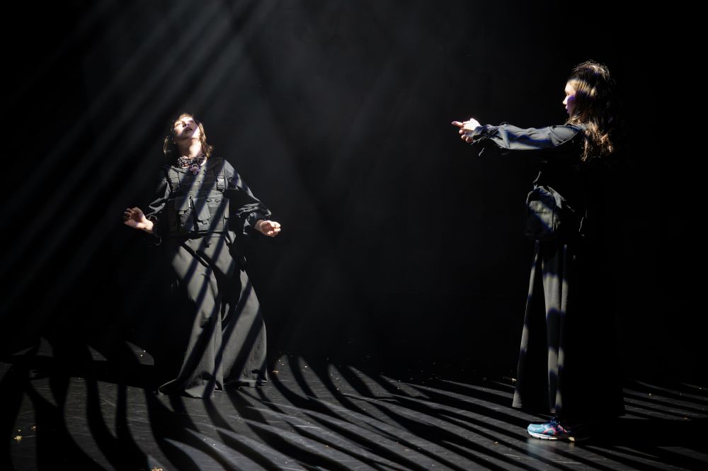 Dwie osoby w długich czarnych szatach stoją na ciemnej scenie, a dramatyczne promienie światła przecinają teatralną atmosferę. Jedna z nich odchyla się do tyłu z uniesionymi ramionami, podczas gdy druga wskazuje, tworząc pełną napięcia scenę, która bada istnienie w tym tajemniczym miejscu.