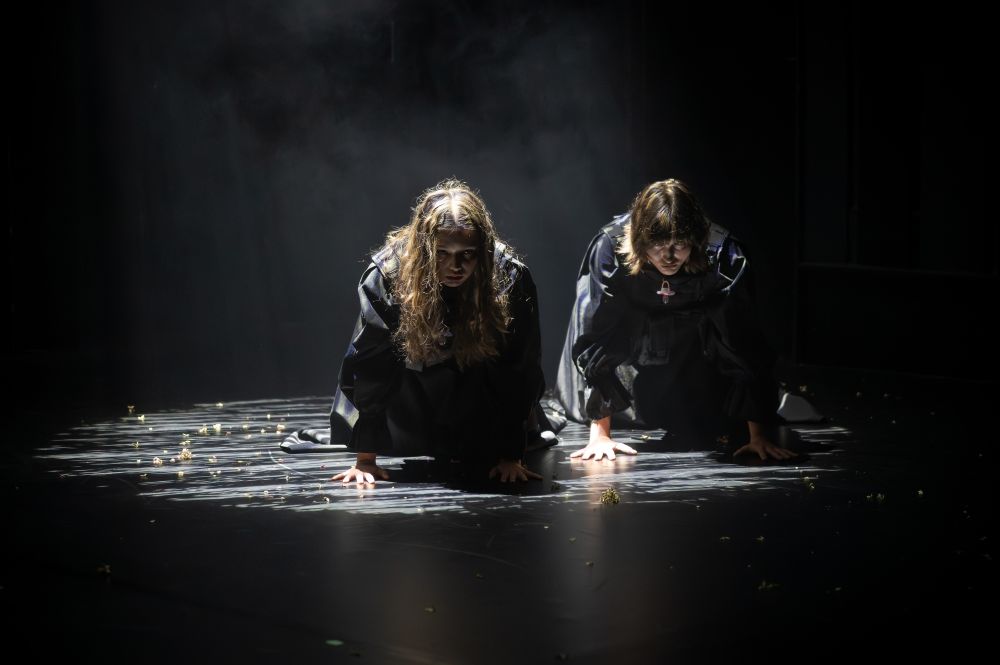 Dwie osoby w ciemnych ubraniach kucają na słabo oświetlonej scenie w teatrze, z rękami na podłodze i opuszczonymi głowami. Dramatyczne oświetlenie rzuca cienie na rozrzucone gruzy, tworząc intensywne i teatralne miejsce.