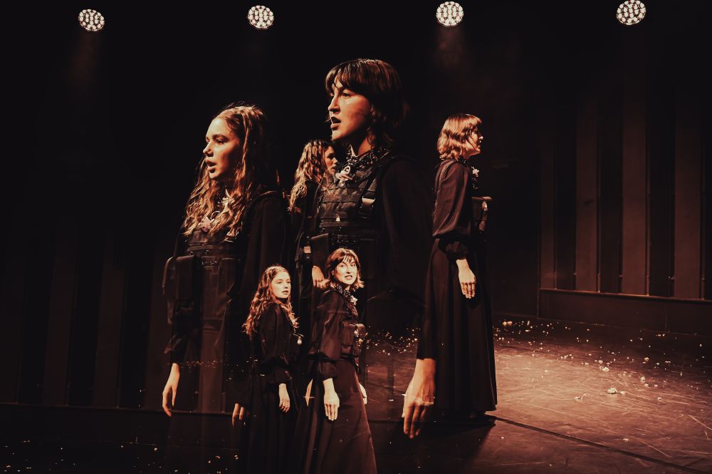 Trzy kobiety w czarnych sukienkach stoją na słabo oświetlonej scenie teatralnej z dramatycznym oświetleniem. Obraz zawiera efekt podwójnej ekspozycji, tworząc upiorne, nakładające się na siebie duplikaty. Rozrzucone gruzy pokrywają miejsce, w którym występują.