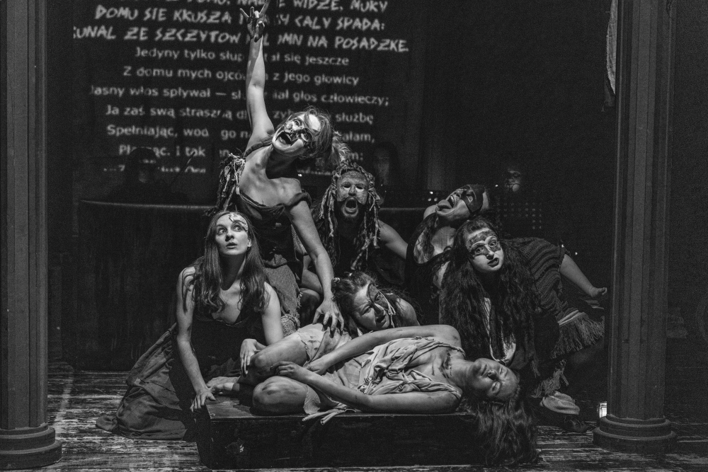 Siedmiu wykonawców Teatru KTO w dramatycznym makijażu i kostiumach pozuje ekspresyjnie na scenie; niektórzy stoją, inni kucają lub leżą, z intensywną mimiką twarzy. Na ścianie za nimi wyświetlany jest tekst. Czarno-biały obraz oddaje żywą energię teatru.