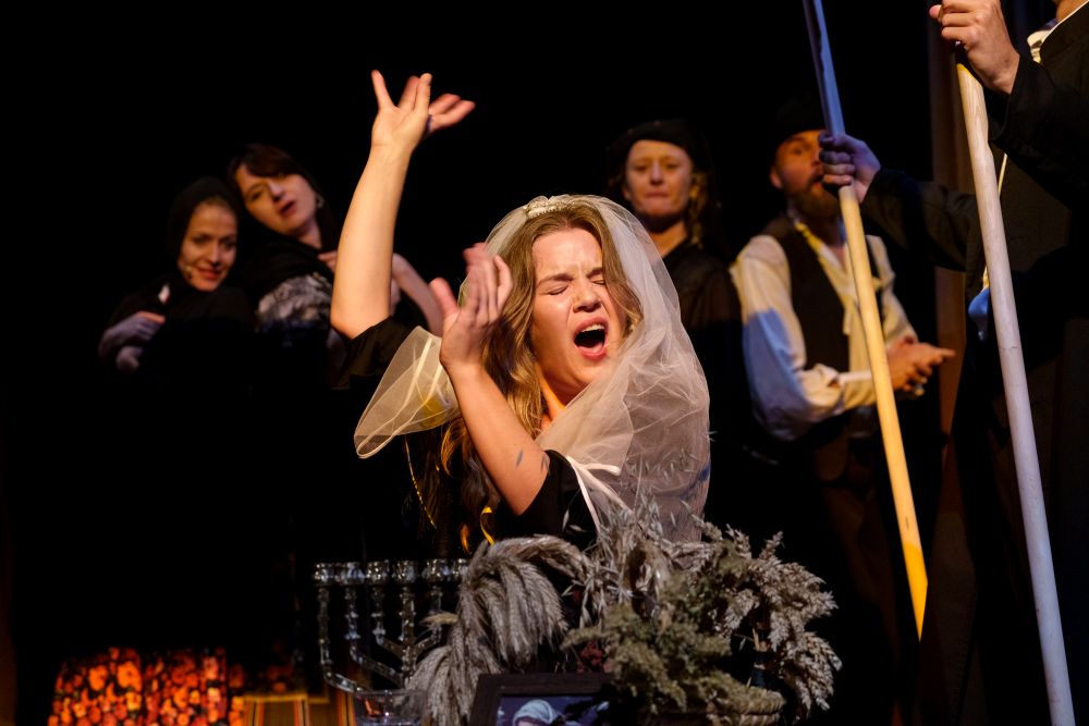 Kobieta w białym welonie unosi ramiona i śpiewa namiętnie na scenie, otoczona innymi aktorami w ciemnych strojach, tworząc dramatyczną scenę teatralną przypominającą sugestywny wieczór teatralny w Teatrze KTO.