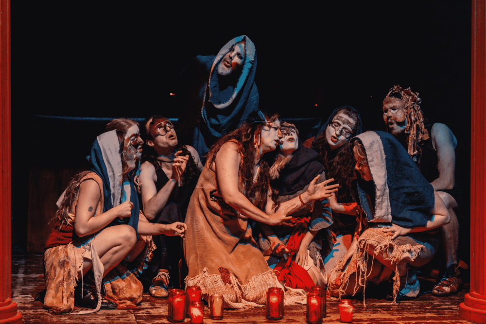 Grupa aktorów z Teatru KTO w podartych kostiumach i upiornie pomalowanych twarzach gromadzi się na scenie, niektórzy klęczą, inni stoją, oświetleni czerwonymi świecami na podłodze, tworząc dramatyczną i tajemniczą scenę teatralną.