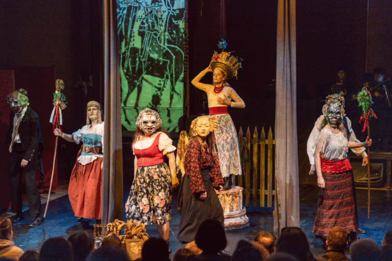 Sześciu wykonawców z Gardzienic stoi na scenie teatralnej w wyszukanych kostiumach i maskach, oświetlonych dramatycznym światłem. Jeden z nich, ukoronowany i uniesiony, utrzymuje pozę, podczas gdy inni w tradycyjnych strojach z laskami występują przed uważną publicznością.