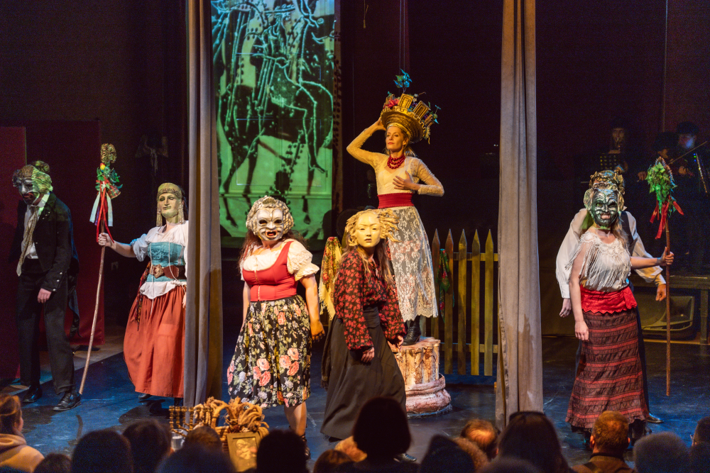 Sześciu wykonawców z Gardzienic stoi na scenie teatralnej w wyszukanych kostiumach i maskach, oświetlonych dramatycznym światłem. Jeden z nich, ukoronowany i uniesiony, utrzymuje pozę, podczas gdy inni w tradycyjnych strojach z laskami występują przed uważną publicznością.