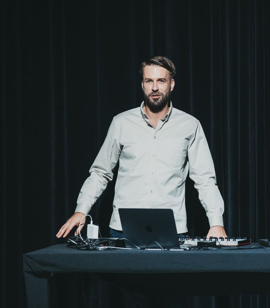 Mężczyzna z brodą stoi za stołem z laptopem, kablami i sprzętem audio, przed czarnymi zasłonami, ubrany w jasną koszulę i patrzy w kamerę.