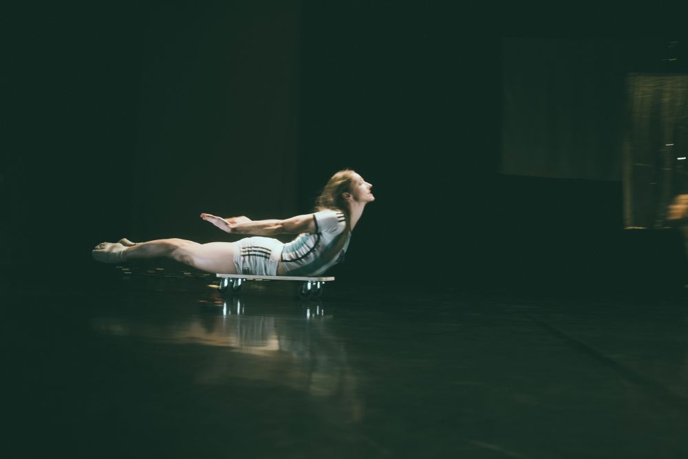 Osoba w białym stroju leży twarzą w dół, z ramionami wyciągniętymi do tyłu, balansując na niskiej platformie na kółkach, gdy przesuwa się po ciemnej scenie Spektaklu - uderzający moment z TrueMoms Show.