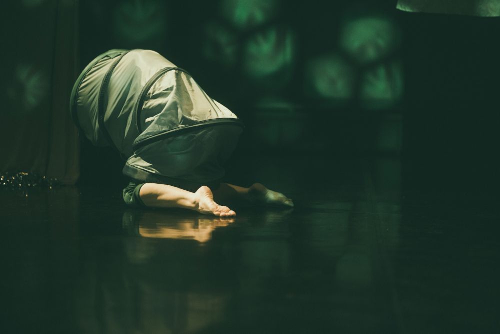 Osoba ubrana w luźną, jasną odzież jest pochylona na ciemnej, odbijającej światło podłodze podczas Spektakle TrueMoms Show, jej głowa i górna część ciała są zakryte, a bose stopy widoczne. Zielonkawe oświetlenie tworzy abstrakcyjne kształty w tle.