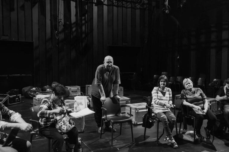 Mężczyzna stoi i opiera się o krzesło w centrum grupy, podczas gdy inni siedzą i rozmawiają lub przeglądają dokumenty w słabo oświetlonym miejscu za kulisami, za którymi stoją skrzynie ze sprzętem - uchwycenie esencji edycji programu KTO?Senior! w czerni i bieli.