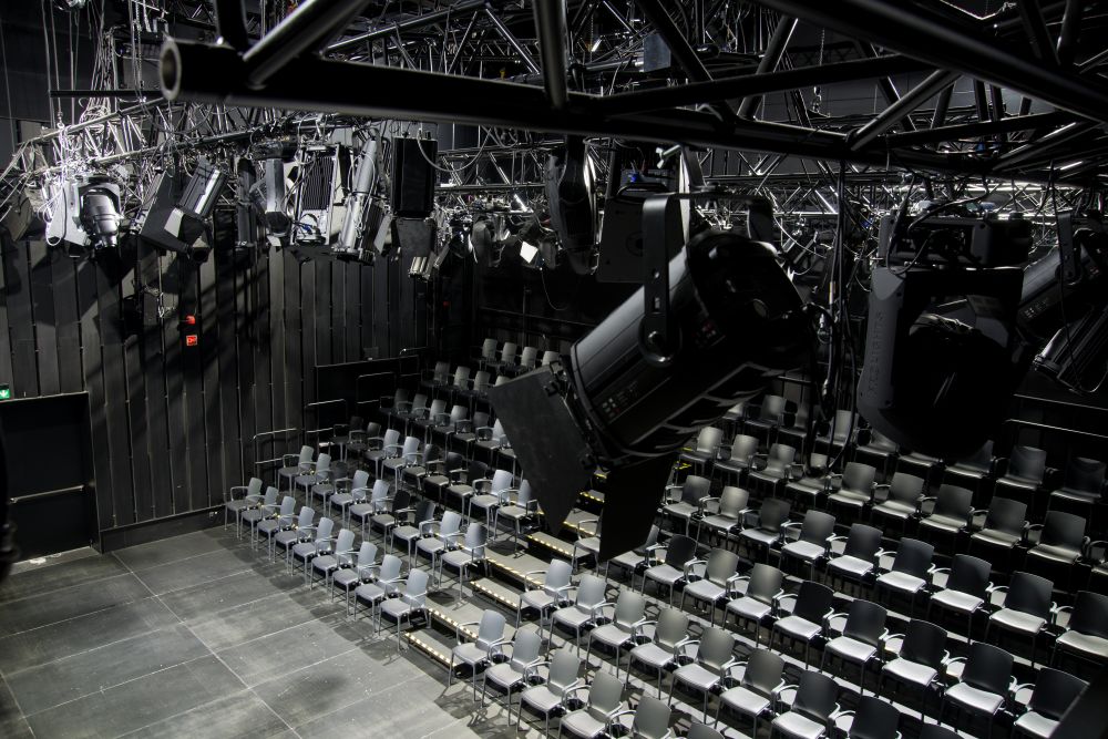 Nowoczesny teatr typu black-box w Teatrze KTO z pustymi szarymi krzesłami ustawionymi w rzędach, otoczony czarnymi ścianami. Liczne światła sceniczne i olinowanie zwisają z sufitu nad miejscami siedzącymi, a pętla indukcyjna zapewnia dostępność dla wszystkich.