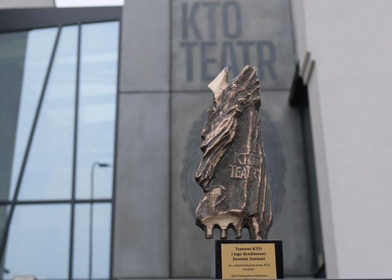 Rzeźba z brązu upamiętniająca Teatr KTO i Jerzego Zonia stoi na czarnym cokole przed nowoczesnym budynkiem, na którym widnieje duży napis "KTO TEATR". Tablica zawiera polską dedykację i odnosi się do Izby Przemysłowo-Handlowej Kraków.