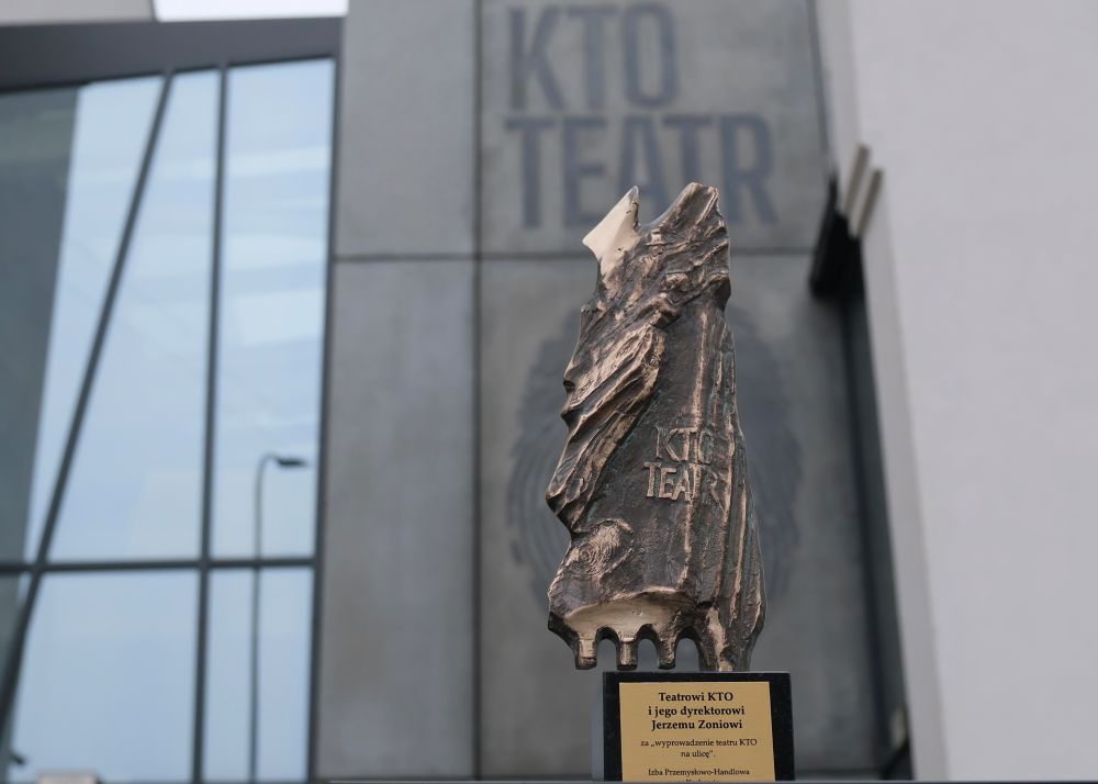 Rzeźba z brązu upamiętniająca Teatr KTO i Jerzego Zonia stoi na czarnym cokole przed nowoczesnym budynkiem, na którym widnieje duży napis "KTO TEATR". Tablica zawiera polską dedykację i odnosi się do Izby Przemysłowo-Handlowej Kraków.
