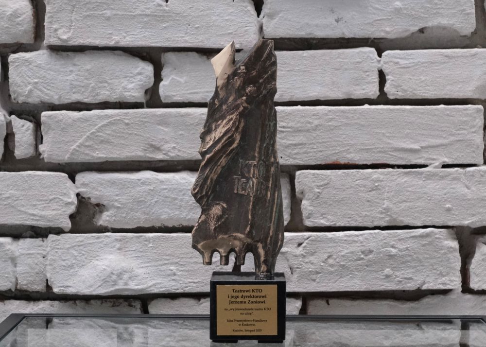 Rzeźbiona metalowa nagroda o abstrakcyjnym kształcie stoi na czarnej prostokątnej podstawie ze złotą tabliczką, na tle białej ceglanej ściany - wyróżnienie przyznane przez Izbę Przemysłowo-Handlową Kraków Jerzemu Zoniowi z Teatru KTO.