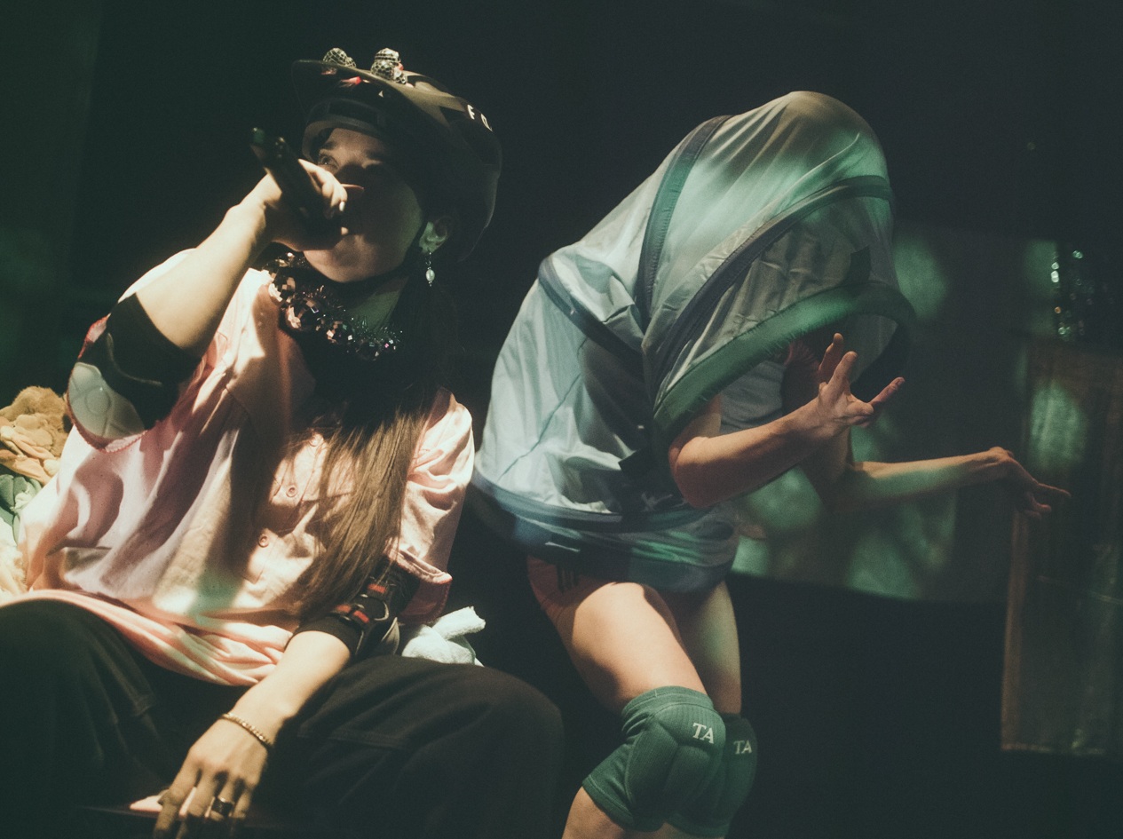 Dwóch wykonawców na scenie: jeden siedzi z mikrofonem w kasku i różowej koszuli, podczas gdy drugi, w szortach i nakolannikach, dramatycznie pozuje pod prześwitującym materiałem - uderzający moment z wyjątkowego pokazu Spektakle.