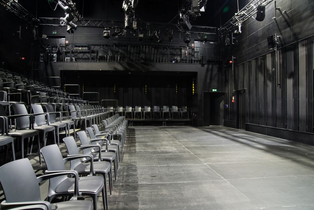 Rzędy pustych, szarych krzeseł w nowoczesnym teatrze czarnej skrzynki Teatru KTO, zwrócone w stronę nagiej sceny pod reflektorami i olinowaniem. Przestrzeń ma czarne ściany, industrialny, minimalistyczny design i jest wyposażona w pętlę indukcyjną dla ułatwienia dostępu.