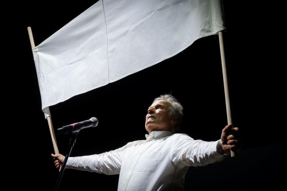 Mężczyzna w białej koszuli stoi na scenie, trzymając dużą białą flagę obiema rękami przy mikrofonie, występując na Międzyuczelnianym Przeglądzie Performans na ciemnym tle.