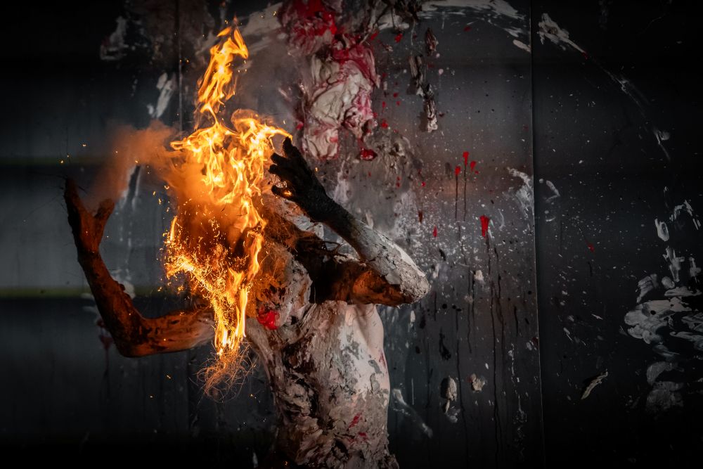 Osoba pokryta białą i czarną farbą stoi na ciemnym, zachlapanym tle, z jaskrawymi pomarańczowymi płomieniami pochłaniającymi jej głowę - dramatyczna scena inspirowana Międzyuczelnianym Przeglądem Performans i aKTOmaR.