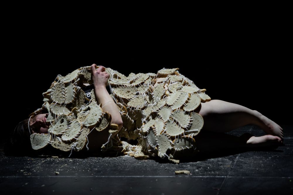 Osoba leży skulona na ciemnej scenie, częściowo przykryta tkanymi lub szydełkowanymi płaskimi przedmiotami przypominającymi chleb lub krakersy, tworząc abstrakcyjną scenę z dramatycznym oświetleniem - uderzający moment z Międzyuczelnianego Przeglądu Performans.